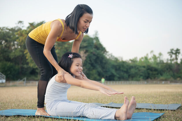 草地妈妈和女儿在做瑜伽在公园里训练妇女和孩子户外运动健康的运动生活方式 坐在帕希莫塔纳萨纳练习 坐在前面弯腰姿势健身伸展瑜伽