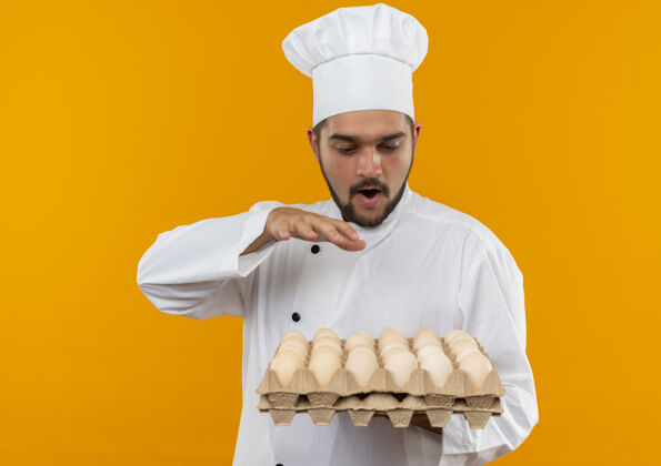 空气穿着厨师制服的年轻男厨师手拿鸡蛋看着一盒一盒的鸡蛋 手放在隔离的橙色墙上印象纸箱烹饪