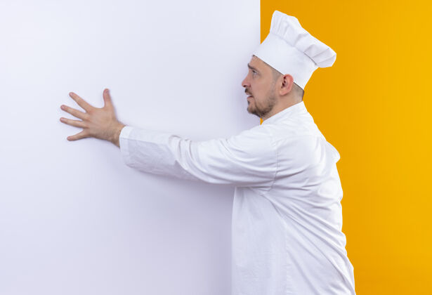 墙年轻帅气的厨师穿着厨师制服站在白色的墙前 把手放在上面 看着橙色墙上孤立的一面印象壁板厨师