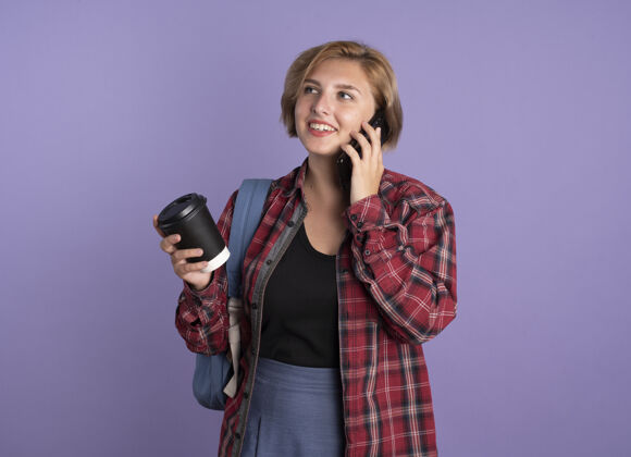 复制带着微笑的年轻斯拉夫学生女孩背着书包拿着纸杯在电话里说话看着身边紫色斯拉夫电话