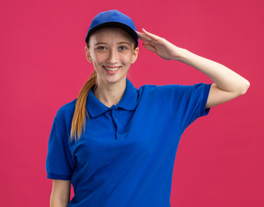 女孩身穿蓝色制服 头戴鸭舌帽的年轻送货员站在粉红色的墙上微笑着自信地敬礼年轻微笑帽子