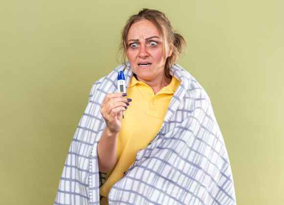 感觉不健康的女人裹在毯子里 感觉很可怕 患流感 感冒 拿着温度计站在绿色的墙上 看起来很害怕害怕流感不健康