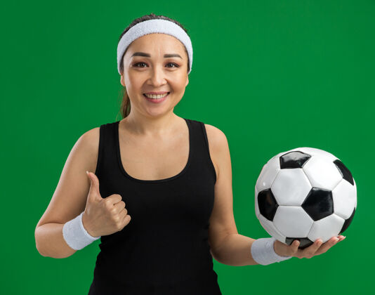 女人戴着头巾 面带微笑地拿着足球站在绿墙上的年轻健身女士年轻站健身