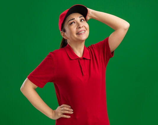 制服身穿红色制服 戴着帽子的年轻女送货员抬起头来 困惑的表情抚摸着站在绿色墙壁上的头帽子立场触摸