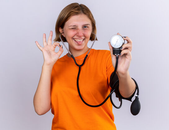 不健康快乐不健康的年轻女子穿着橙色t恤 用眼压计测量血压 脸上带着微笑 站在白墙上做着“ok”的手势使用站立年轻