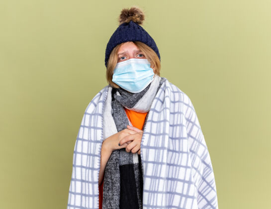 包裹不健康的年轻女子戴着暖和的帽子 裹着毛毯 戴着护面面具 站在绿色的墙上感觉不舒服 患流感和感冒不适年轻女人