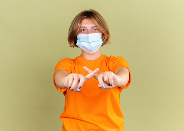 手势不健康的年轻女子穿着橙色t恤 戴着护面面具 感觉不适 感染病毒 停止手势 食指交叉 站在绿色的墙上站立痛苦病毒