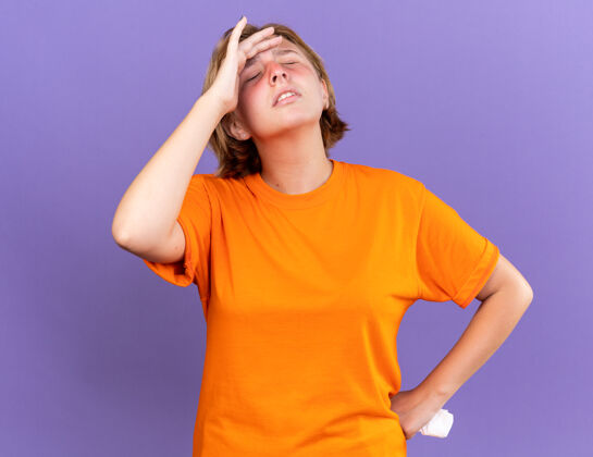 触摸不健康的年轻女子穿着橘色t恤 站在紫色的墙上摸额头感到不适 头晕 感冒同时年轻不适