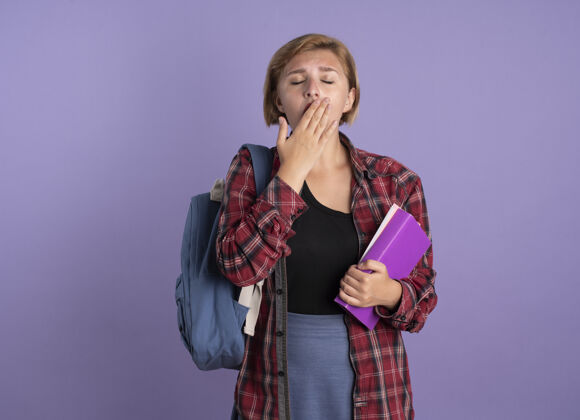 穿昏昏欲睡的年轻斯拉夫女学生 背着背包 手放在嘴上 手里拿着书和笔记本嘴紫色背包