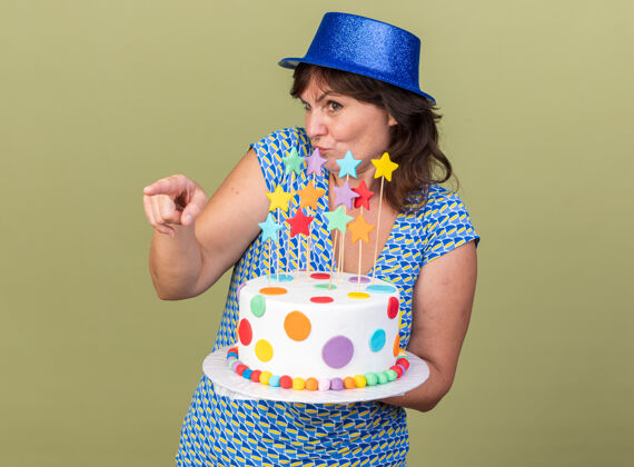 指向困惑的中年妇女戴着派对帽 手里拿着生日蛋糕 一边看一边用食指指着旁边 站在绿色的墙上庆祝生日派对壁板举行蛋糕