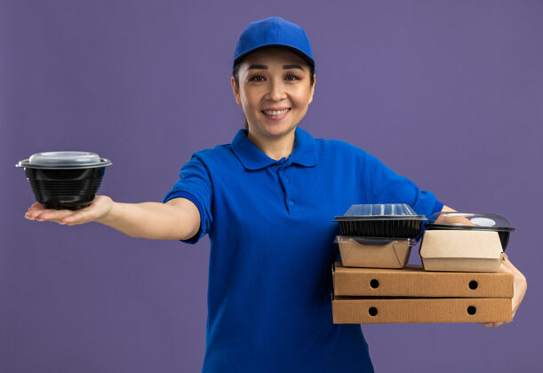 微笑身着蓝色制服 头戴鸭舌帽 手持披萨盒和食品包的快乐年轻送货员站在紫色的墙上 开心地微笑着欢呼站着抱着