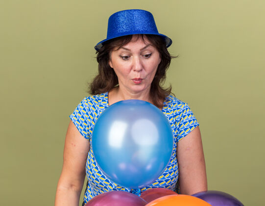 女人中年妇女戴着派对帽 手里拿着一堆五颜六色的气球 站在绿色的墙壁上庆祝生日 看起来既惊讶又惊讶帽子庆祝五颜六色