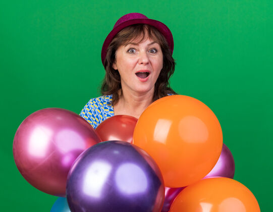 年龄中年妇女戴着派对帽 手持五颜六色的气球 快乐而兴奋地微笑着 站在绿色的墙上欢庆生日派对五颜六色微笑兴奋