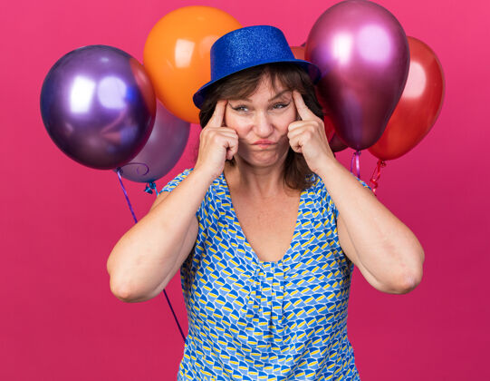 角落中年妇女戴着派对帽 手里拿着五颜六色的气球 把眼角拉向两边 困惑不安地站在粉色的墙上庆祝生日派对气球不高兴拉