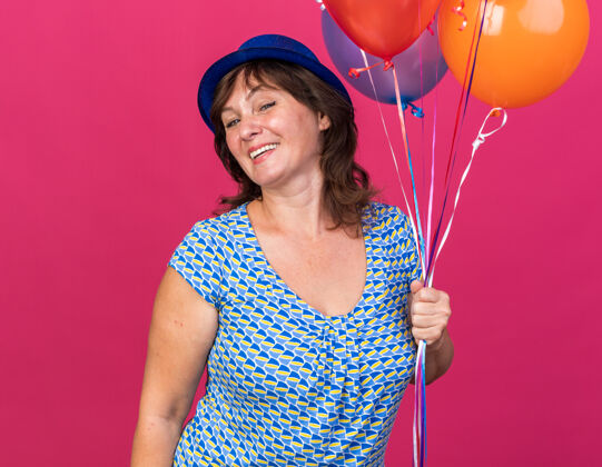 中快乐快乐的中年妇女戴着派对帽 手持一束五颜六色的气球 微笑着站在粉色的墙上庆祝生日派对微笑年龄庆祝