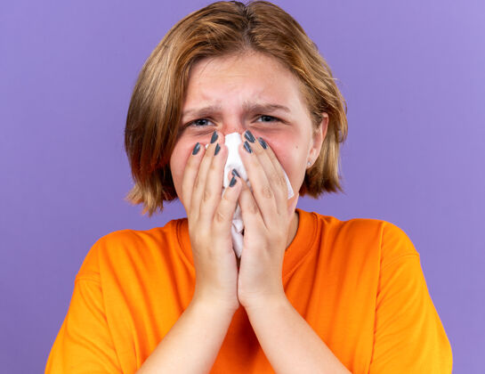 年轻不健康的年轻女子穿着橙色t恤 感觉很糟糕 流鼻涕感冒了 站在紫色的墙上打喷嚏抓到可怕感冒