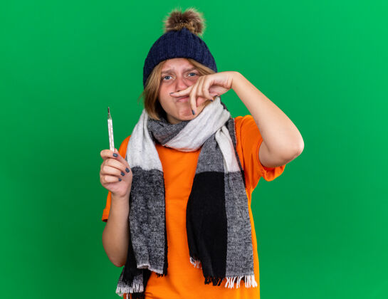 女人不健康的年轻女子穿着橙色t恤 戴着帽子 脖子上围着暖和的围巾 拿着体温计 发烧 鼻子流鼻涕 站在绿色的墙上可怕感觉鼻子