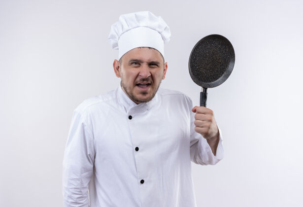 烹饪愤怒的年轻帅哥厨师身着厨师制服 手拿煎锅孤立在白墙上厨师拿着年轻