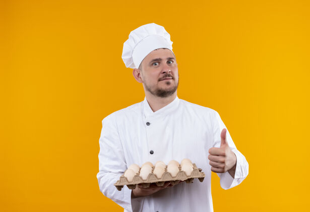烹饪年轻帅气的厨师穿着厨师制服 拿着一盒鸡蛋 在橙色的墙上孤立地竖起大拇指显示大拇指年轻