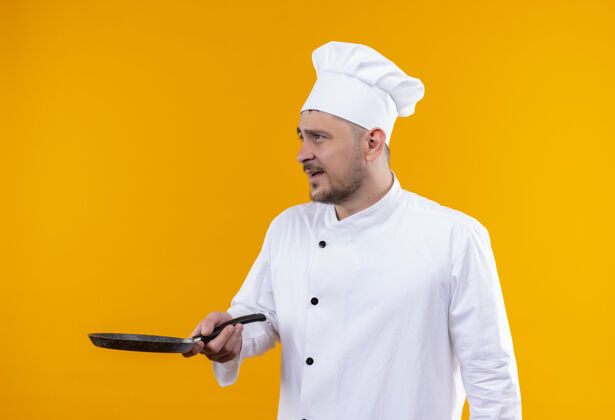 烹饪印象深刻的年轻帅哥厨师穿着厨师制服拿着煎锅 看着隔离在橙色墙上的一面壁板锅薯条