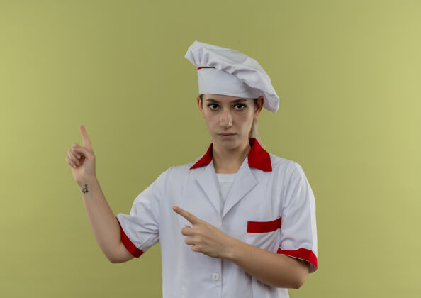 漂亮自信的年轻漂亮厨师 身着厨师制服 指着隔离在绿墙上的一侧指点厨师烹饪