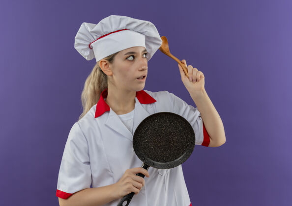 壁板年轻漂亮的厨师穿着厨师制服 手里拿着煎锅和勺子 看着紫色墙上孤立的一面持有烹饪薯条
