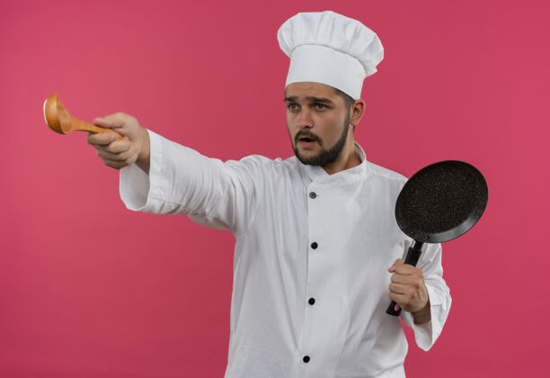 勺子穿着厨师制服的年轻男厨师手拿煎锅 伸出勺子 看着粉色墙壁上孤立的一面印象制服薯条