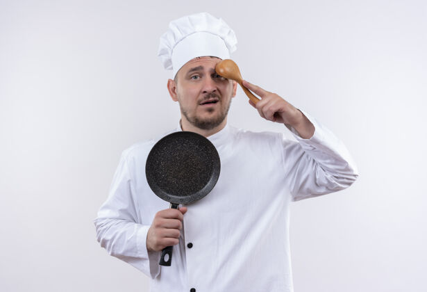 厨师困惑的年轻帅哥厨师穿着厨师制服拿着煎锅 把勺子放在白色墙壁上孤立的额头上帅气年轻制服