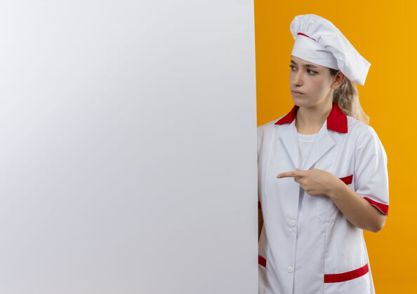 烹饪困惑的年轻漂亮厨师穿着厨师制服站在后面指着和看着白色的墙壁隔离在橙色的墙壁与复制空间站着制服墙