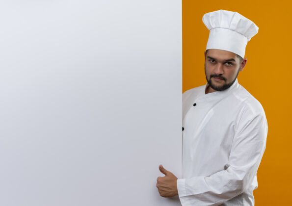 烹饪身着厨师制服的自信年轻男厨师站在白色墙壁后 竖起大拇指 看起来孤立地站在橙色墙壁上 留有复印空间展示自信站立