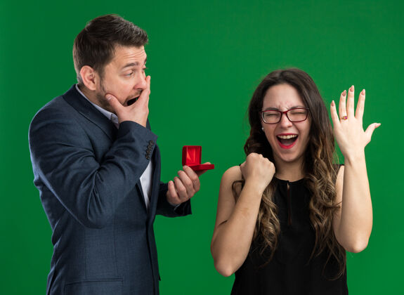 困惑年轻漂亮的夫妇困惑的男人拿着红盒子向他可爱兴奋的女友求婚 手指上戴着订婚戒指站在绿色的墙上庆祝情人节年轻举行求婚