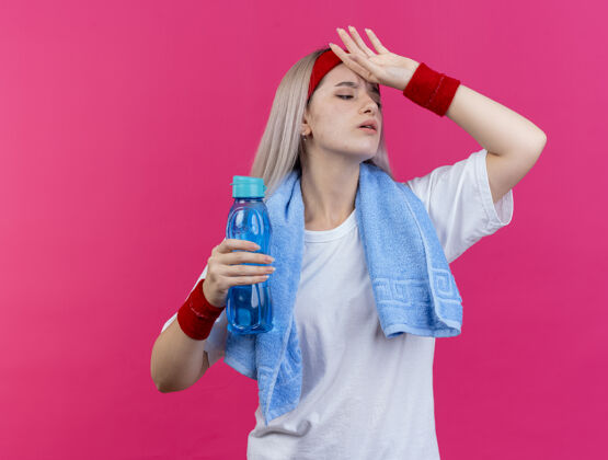 头带疼痛的年轻白人运动女孩戴着背带 脖子上戴着毛巾 戴着头带和腕带 手放在额头上 拿着水瓶毛巾粉红色瓶子