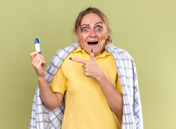 痛苦不健康的女人裹着毯子 感觉很可怕 患了流感和感冒 拿着温度计 用食指指着它 站在绿色的墙上 看起来很害怕目录手指感冒