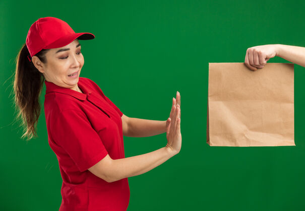 帽子身穿红色制服 戴着帽子的年轻送货女孩站在绿色的墙上拒绝接受纸质包裹包装拒绝女孩