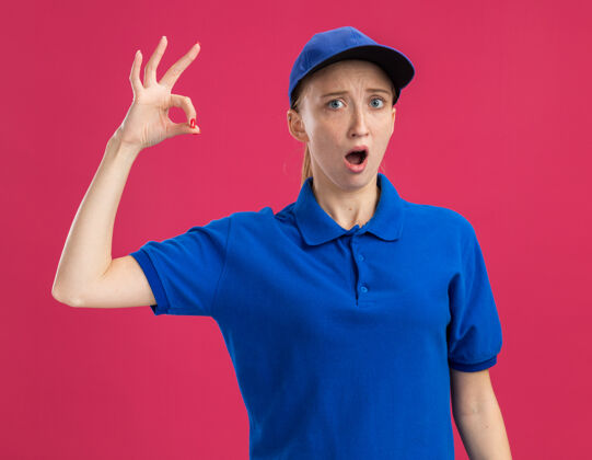 年轻身穿蓝色制服 头戴鸭舌帽的年轻送货员惊讶地在粉红色的墙上立着“ok”的牌子惊喜送货表演