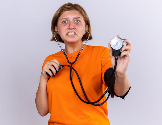 不健康不健康的年轻女子穿着橙色t恤 感觉不舒服 站在白色的墙上用血压计测量血压 看起来很担心和失望担心失望女人