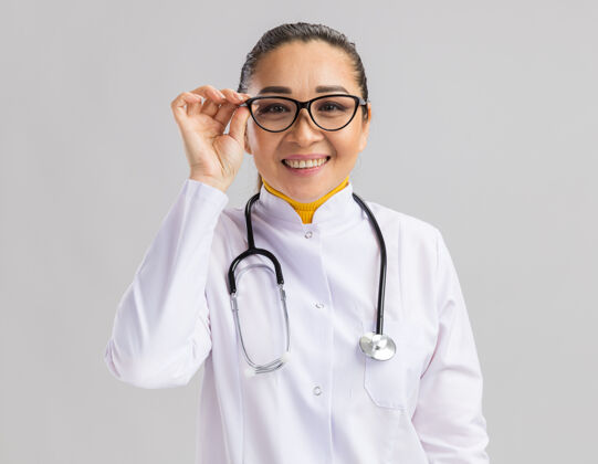 微笑快乐的年轻女医生 穿着白大褂 戴着眼镜 脖子上戴着听诊器 脸上带着微笑 站在白墙上医生戴着女人