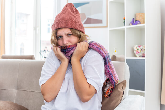 不健康不健康的年轻女子戴着暖和的帽子 脖子上围着围巾 感觉不舒服 生病 患感冒和流感 看起来很担心 坐在客厅里的沙发上年轻房间暖和