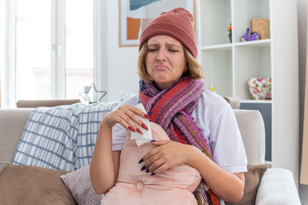 不健康不健康的年轻女士戴着暖和的帽子 戴着围巾 看起来不舒服 生病了 拿着纸巾 患感冒和流感 还擤鼻涕 坐在明亮的客厅沙发上吹房间鼻子