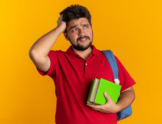 困惑年轻的留着胡子的学生 穿着红色马球衫 背着背包 拿着笔记本 一边看着一边 一边困惑地用手放在头上 因为他站错了笔记本手背包