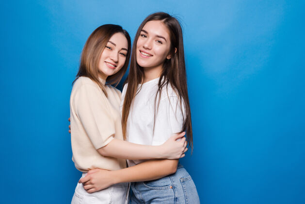 微笑两个女朋友拥抱着彼此孤立的蓝色墙壁拥抱年轻快乐