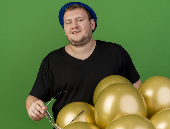 绿色高兴的成年斯拉夫男子手持眼镜戴着蓝色派对帽站在氦气球穿氦举行