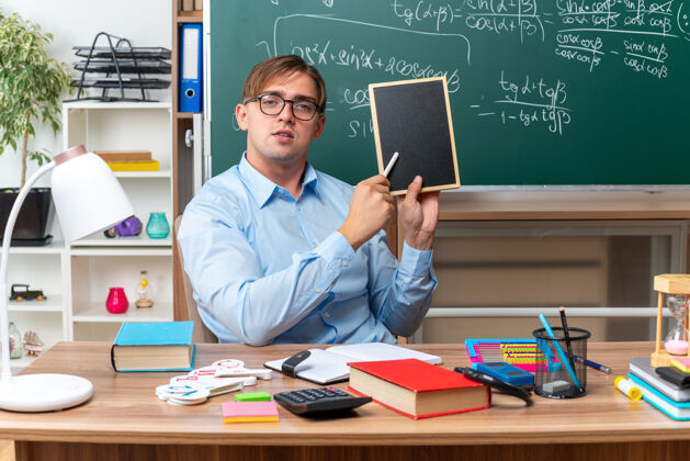 男年轻的男老师戴着眼镜 用小黑板和粉笔讲解课文 自信地坐在课桌旁 教室里黑板前放着书和笔记人课自信
