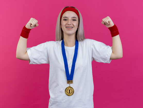 运动微笑的年轻白人运动女孩 戴着牙套 脖子上戴着金牌 戴着头带和腕带 紧张的二头肌头带背带微笑