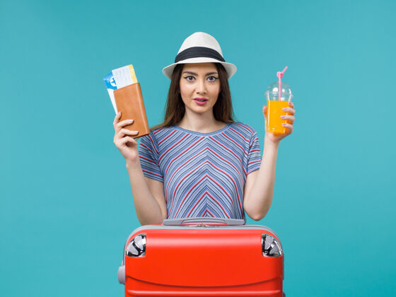 人正面图：度假中的女人拿着她的果汁和蓝色背景的机票去度假举行票旅行