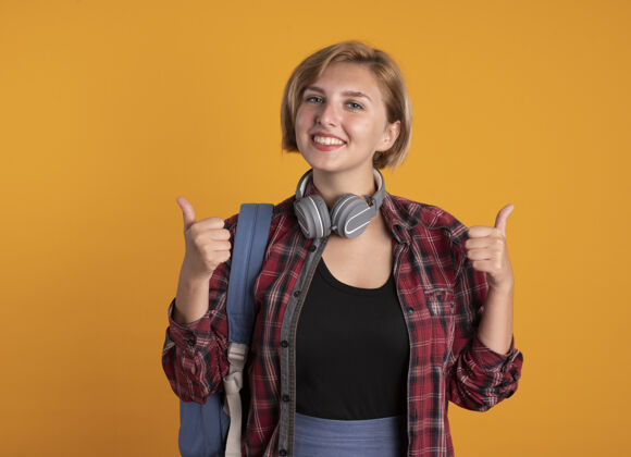 橙色微笑的年轻斯拉夫女学生戴着耳机背着背包双手竖起大拇指斯拉夫背包手