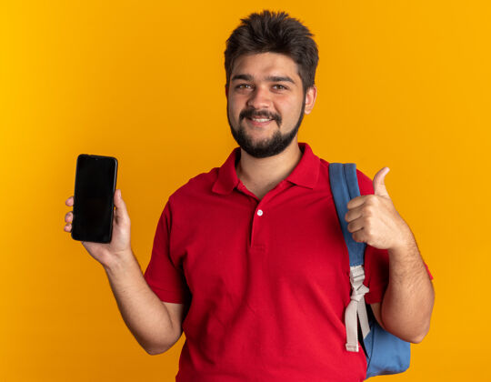马球年轻的留着胡子的学生 穿着红色马球衫 背着背包 拿着智能手机 微笑着 自信地竖起大拇指站着胡须站着背包