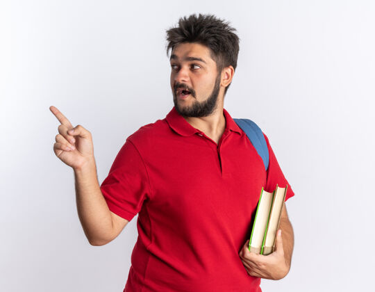靠边年轻的留着胡子的学生 穿着红色马球衫 背着背包 拿着笔记本 看着一边 用食指指着一边微笑着站着红色站着指着
