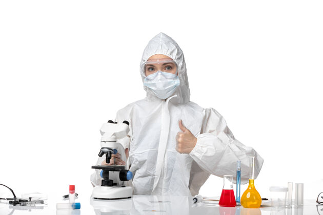 显微镜正面图年轻女医生穿着白色防护服 戴着口罩 由于科维德用显微镜在浅白色背景上大流行 溅起健康的科维德大流行专业套装