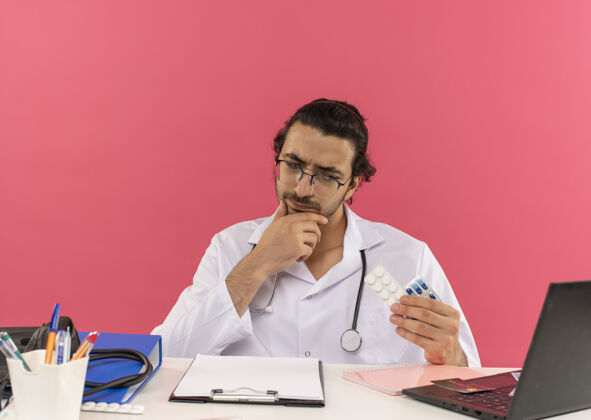 复制年轻的男医生戴着医用眼镜 穿着医用长袍 听诊器坐在办公桌旁思考穿坐